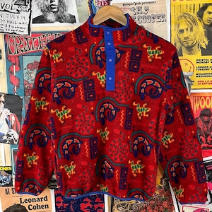 May include: A red fleece pullover featuring a pattern of blue, green, and yellow shapes. The collar and placket are blue, with three buttons. The garment is displayed against a backdrop of vintage posters.