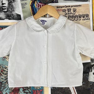 May include: A white, short-sleeved blouse with a Peter Pan collar. The blouse has three buttons down the front and is hanging on a wooden hanger. The collar has a small blue and white tag. The blouse is displayed against a backdrop of vintage posters.