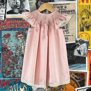 May include: A light pink baby dress with a smocked bodice and ruffled sleeves. The dress has a dotted pattern and white lace trim. The dress is hanging on a wooden hanger against a backdrop of vintage concert posters.