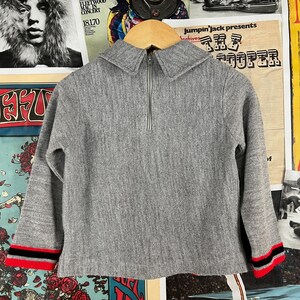 Puede incluir: Un su&eacute;ter gris de manga larga con una cremallera en la parte delantera. El su&eacute;ter tiene un cuello y rayas rojas y negras en los pu&ntilde;os.