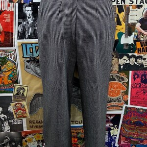 Pode incluir: Cal&ccedil;a xadrez preta e branca com pregas. A cal&ccedil;a est&aacute; em um manequim e &eacute; exibida contra um fundo de cartazes de concertos vintage.