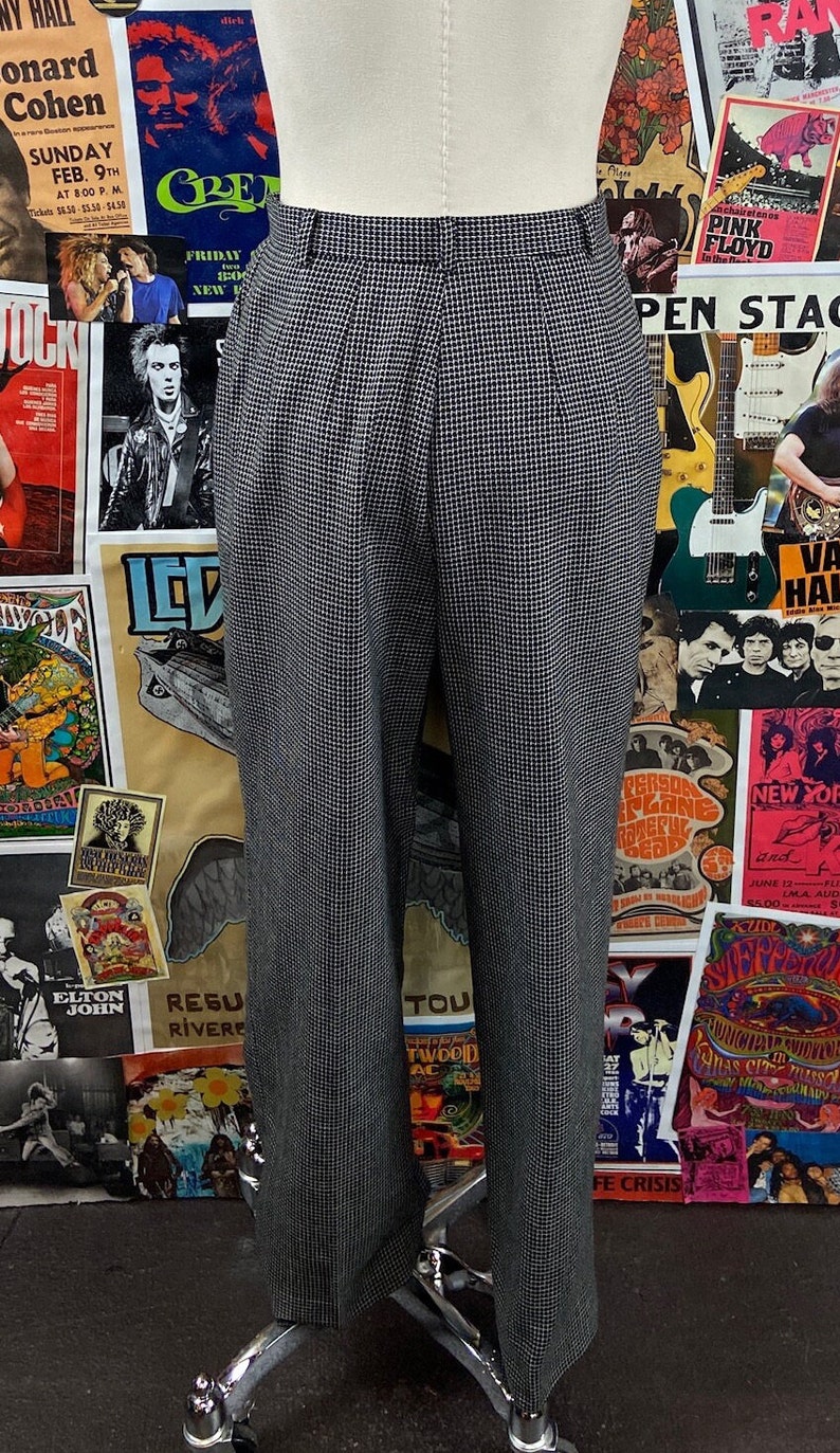 Pode incluir: Cal&ccedil;a xadrez preta e branca com pregas. A cal&ccedil;a est&aacute; em um manequim e &eacute; exibida contra um fundo de cartazes de concertos vintage.