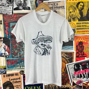 May include: White t-shirt featuring a black graphic of a man in a sombrero with the text "ACAPULCO GOLD". The shirt is hanging on a wooden hanger against a backdrop of vintage concert posters. The shirt's design is centered on the front.