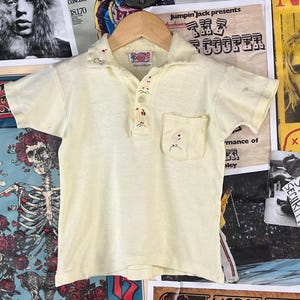 May include: Cream-colored vintage polo shirt with short sleeves, a collar, and a chest pocket. The shirt features embroidered details and a button-down placket. The shirt is displayed against a backdrop of vintage posters.