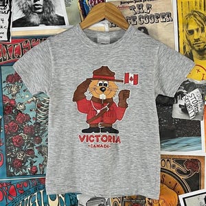 Könnte beinhalten: Ein graues T-Shirt mit einem Cartoon-Biber in roter Uniform, der eine kanadische Flagge hält. Der Biber trägt einen braunen Hut und salutiert. Der Text "VICTORIA CANADA" ist darunter gedruckt.
