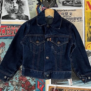 May include: A dark blue denim jacket with a classic collar, button-up front, and two chest pockets. The jacket has a vintage look with metal buttons and a small orange tag. The jacket is displayed on a wooden hanger against a backdrop of vintage posters.