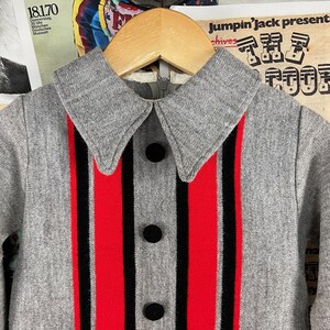 Puede incluir: Un su&eacute;ter gris con cuello y rayas verticales rojas y negras. El su&eacute;ter tiene cierre de botones y un cuello grande.