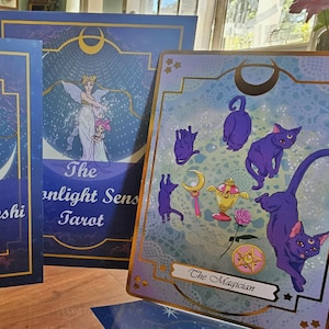 May include: A deck of tarot cards featuring a blue and purple color palette. One card displays the text The Magician with illustrations of purple cats, a golden chalice, a pink rose, and a star emblem. Another card shows The Moonlight Senshi Tarot.