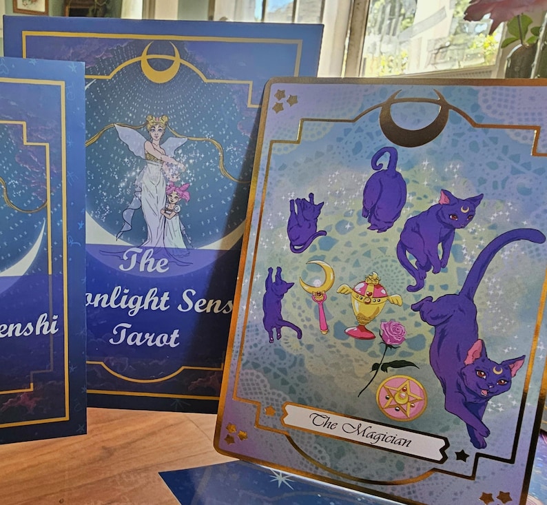 May include: A deck of tarot cards featuring a blue and purple color palette. One card displays the text The Magician with illustrations of purple cats, a golden chalice, a pink rose, and a star emblem. Another card shows The Moonlight Senshi Tarot.
