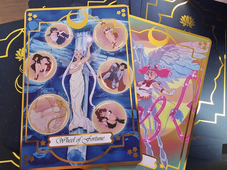 May include: A rectangular tarot card featuring a central figure in a white gown standing before a pillar, surrounded by five circular vignettes of anime characters. The card has a dark blue background with gold borders and the text Wheel of Fortune.