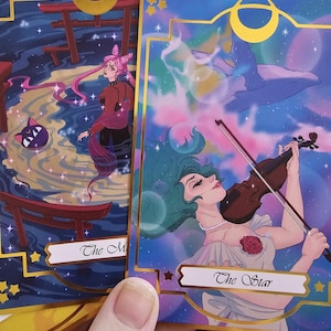 May include: Two rectangular tarot cards with gold foil borders. One card titled The Star features a person with green hair playing a violin against a cosmic background. The other card shows a person with pink hair and a cat in a dark water scene.