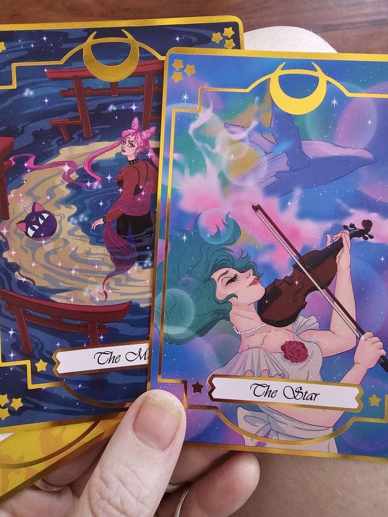 May include: Two rectangular tarot cards with gold foil borders. One card titled The Star features a person with green hair playing a violin against a cosmic background. The other card shows a person with pink hair and a cat in a dark water scene.