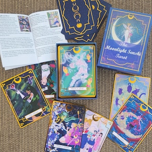 May include: A tarot card deck titled The Moonlight Senshi Tarot, featuring a blue box with gold crescent moon accents, a guidebook, and several illustrated cards depicting anime-style characters, magical wands, and cats with gold-gilded edges.
