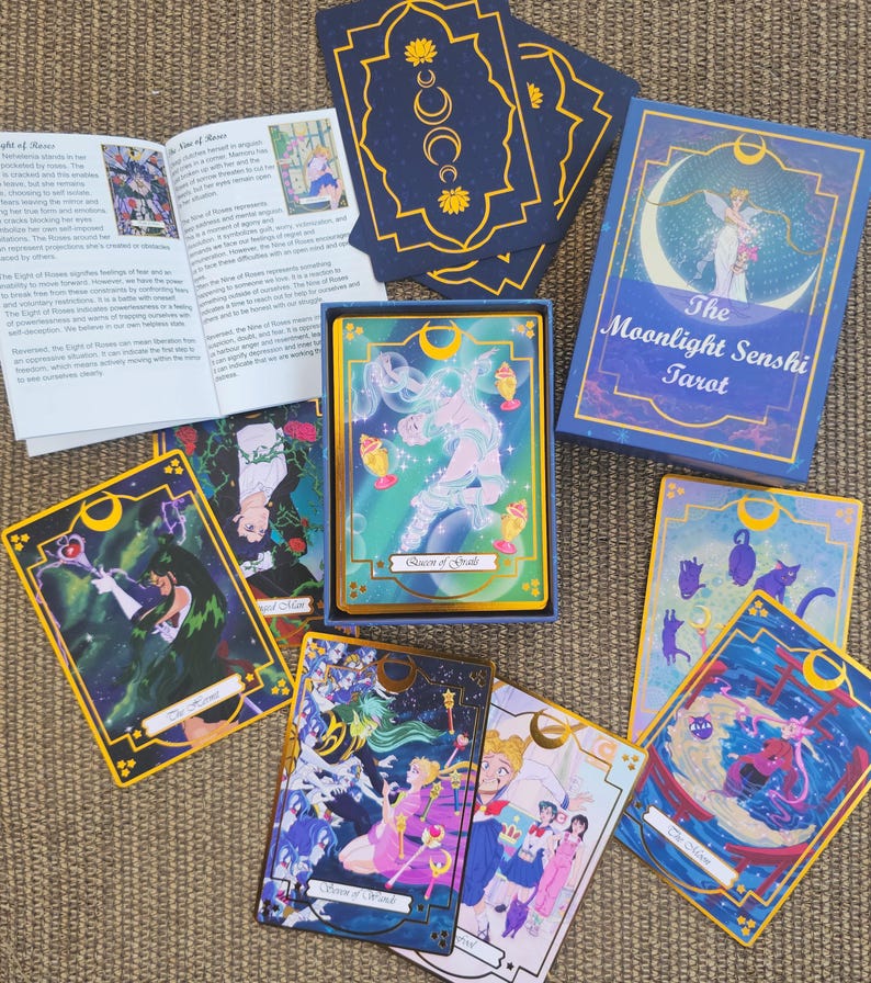May include: A tarot card deck titled The Moonlight Senshi Tarot, featuring a blue box with gold crescent moon accents, a guidebook, and several illustrated cards depicting anime-style characters, magical wands, and cats with gold-gilded edges.