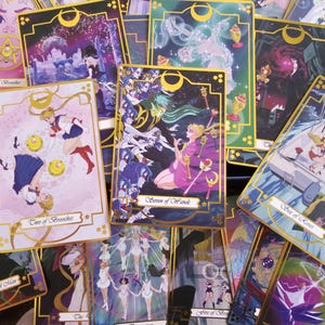 May include: A collection of rectangular tarot cards featuring anime-style illustrations with gold borders and crescent moon motifs. The cards display various characters in colorful scenes, including the Seven of Wands and Two of Brooches, with text labels printed in a cursive script at the bottom of each card.