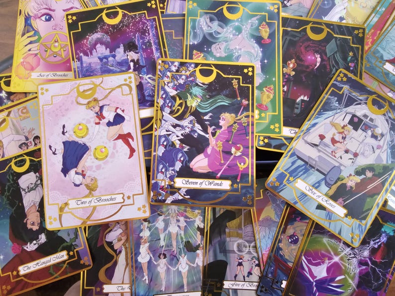 May include: A collection of rectangular tarot cards featuring anime-style illustrations with gold borders and crescent moon motifs. The cards display various characters in colorful scenes, including the Seven of Wands and Two of Brooches, with text labels printed in a cursive script at the bottom of each card.