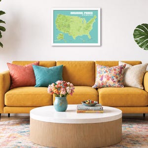 May include: A living room scene featuring a mustard yellow sofa with colorful throw pillows. A framed map of U.S. National Parks hangs above the sofa. A round coffee table holds books, a bowl of snacks, and a vase of flowers. A potted plant is on the left.