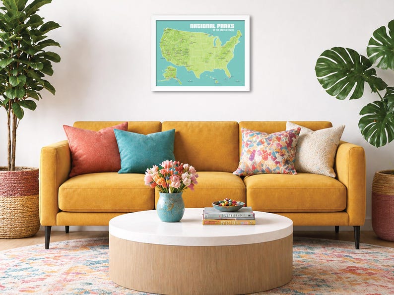 May include: A living room scene featuring a mustard yellow sofa with colorful throw pillows. A framed map of U.S. National Parks hangs above the sofa. A round coffee table holds books, a bowl of snacks, and a vase of flowers. A potted plant is on the left.