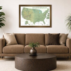 May include: A framed map of the United States National Park System hangs above a brown sofa. The map is in shades of green and brown, with text at the top. A round, dark brown coffee table sits in front of the sofa, with plants on either side.