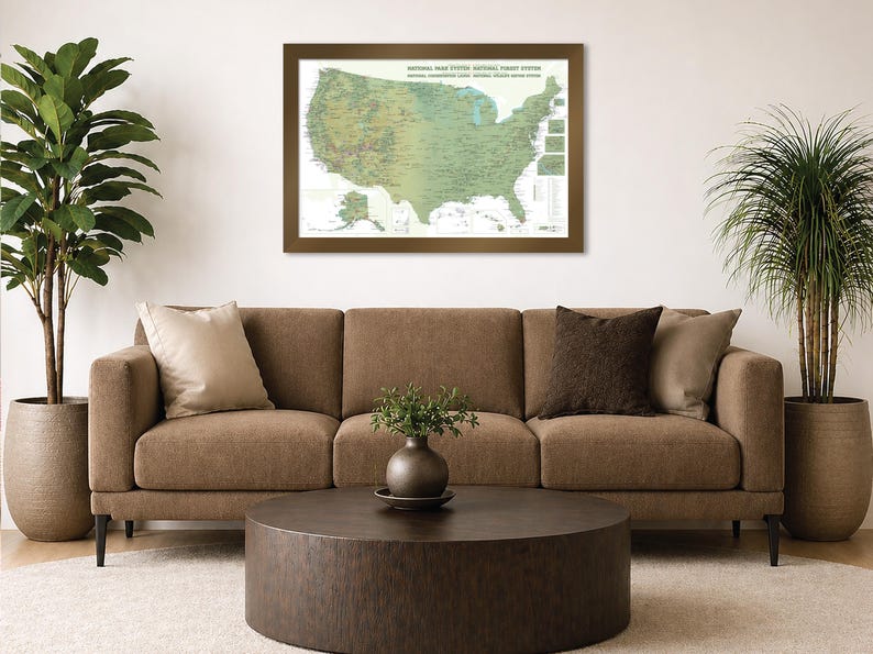 May include: A framed map of the United States National Park System hangs above a brown sofa. The map is in shades of green and brown, with text at the top. A round, dark brown coffee table sits in front of the sofa, with plants on either side.
