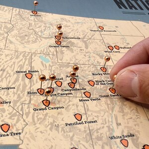 May include: A detailed map of the United States with copper-coloured push pins marking locations. The map highlights national parks with orange shield icons. A hand is placing a pin on the map.