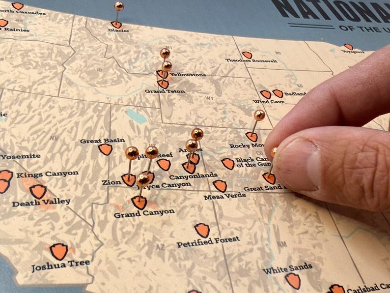 May include: A detailed map of the United States with copper-coloured push pins marking locations. The map highlights national parks with orange shield icons. A hand is placing a pin on the map.