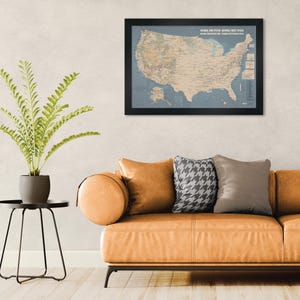 May include: A framed map of the United States, showcasing the National Park System and National Forest System. The map is in a black frame and hangs above a tan leather sofa with decorative pillows. A potted plant sits on a black side table.