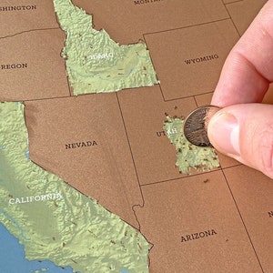 May include: A brown scratch-off map of the United States with state names. A hand holds a coin, scratching off the state of Utah. The map shows the states in a light green color after being scratched.