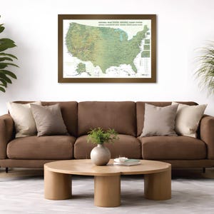 May include: A framed map of the United States National Park System hangs above a brown sofa with decorative pillows. A round wooden coffee table sits in front of the sofa. Potted plants flank the sofa, adding a touch of greenery to the room.