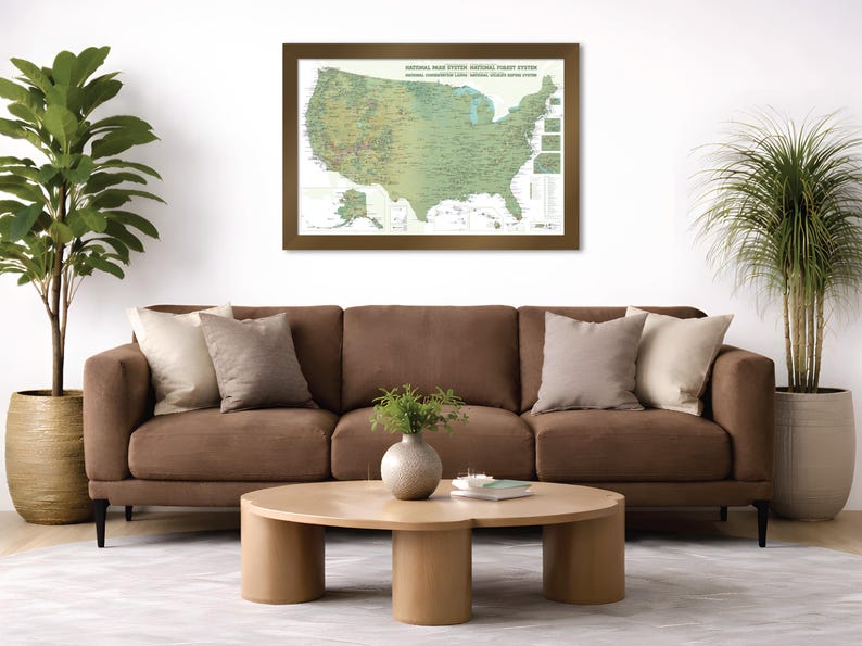 May include: A framed map of the United States National Park System hangs above a brown sofa with decorative pillows. A round wooden coffee table sits in front of the sofa. Potted plants flank the sofa, adding a touch of greenery to the room.