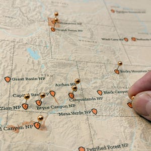 May include: A detailed map of the western United States with push pins marking national parks. The map features names like Zion NP, Grand Canyon NP, and Arches NP. A hand is placing a pin on the map.