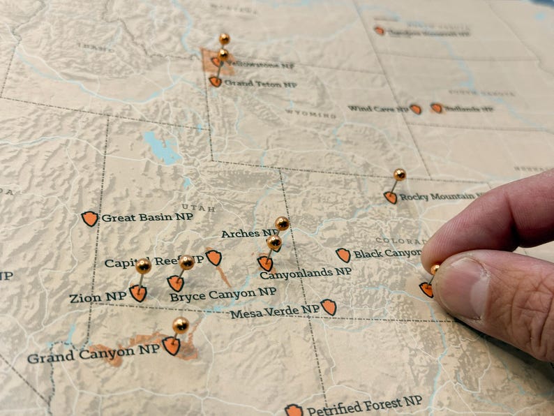 May include: A detailed map of the western United States with push pins marking national parks. The map features names like Zion NP, Grand Canyon NP, and Arches NP. A hand is placing a pin on the map.
