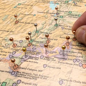 May include: A detailed map of the southwestern United States, with numerous push pins marking locations. The pins are gold, silver, and copper-colored, highlighting various national parks and landmarks. The map is tan and beige.