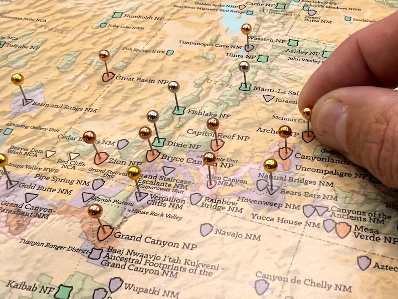 May include: A detailed map of the southwestern United States, with numerous push pins marking locations. The pins are gold, silver, and copper-colored, highlighting various national parks and landmarks. The map is tan and beige.