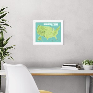 May include: A framed map of the United States National Parks. The map is green on a teal background, with the text "NATIONAL PARKS of the UNITED STATES" at the top. The frame is white. The map is hanging on a light grey wall.