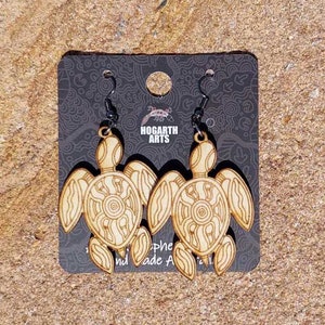 May include: A pair of wooden turtle earrings with intricate designs. The earrings are on a black card with the text "HOGARTH ARTS" printed on it.