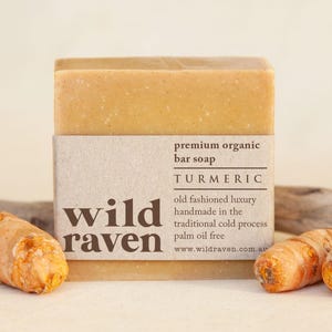 May include: A bar of turmeric soap with a brown paper label that reads "premium organic bar soap TURMERIC old fashioned luxury handmade in the traditional cold process palm oil free www.wildraven.com.au" and the words "wild raven" in bold letters. The soap is next to two pieces of turmeric root.