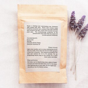 May include: A brown paper bag with a white label containing instructions for a relaxing bath soak. The label lists the ingredients: Epsom salt, sea salt, lavender flower petals, and lavender essential oil. The text on the label reads "Enjoy a relaxing and rejuvenating spa treatment with herbal bath salts. These salts contain many beneficial minerals and nutrients that help detoxify and condition the skin, soothe tired muscles and ease joint pain. The aromatherapy properties of the infused herbs also help relax the mind and increase general wellbeing."