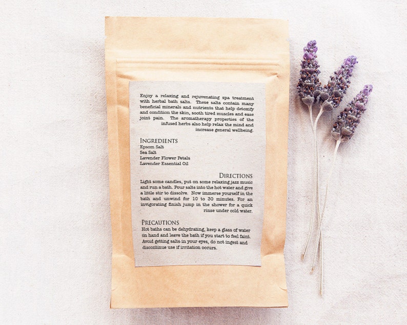 May include: A brown paper bag with a white label containing instructions for a relaxing bath soak. The label lists the ingredients: Epsom salt, sea salt, lavender flower petals, and lavender essential oil. The text on the label reads "Enjoy a relaxing and rejuvenating spa treatment with herbal bath salts. These salts contain many beneficial minerals and nutrients that help detoxify and condition the skin, soothe tired muscles and ease joint pain. The aromatherapy properties of the infused herbs also help relax the mind and increase general wellbeing."