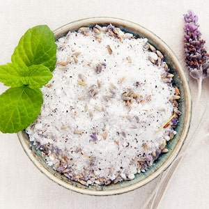 May include: A bowl of white bath salts with lavender buds and a sprig of green mint leaves.