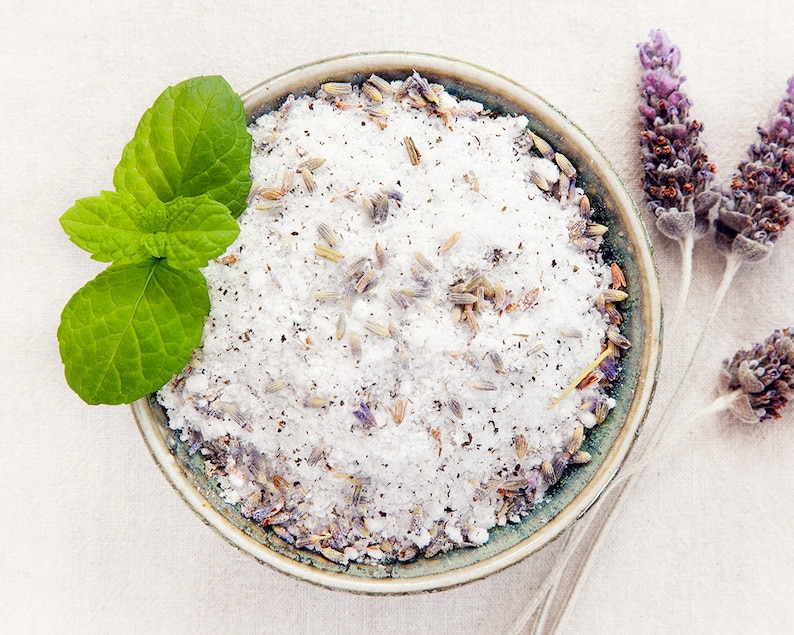 May include: A bowl of white bath salts with lavender buds and a sprig of green mint leaves.