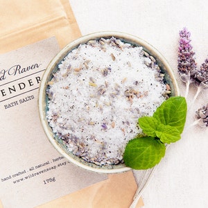 May include: A small bowl filled with white bath salts and lavender buds. The bowl is on a white fabric background with dried lavender sprigs and a sprig of green mint.