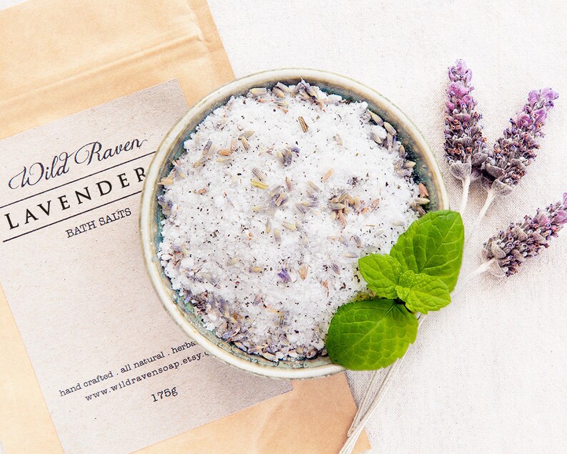 May include: A small bowl filled with white bath salts and lavender buds. The bowl is on a white fabric background with dried lavender sprigs and a sprig of green mint.
