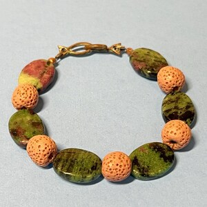 May include: A bracelet with green and red oval stones and coral-coloured spherical beads. The bracelet is fastened with a gold-coloured clasp. The stones and beads are arranged in a circular pattern, creating a unique piece of jewellery.