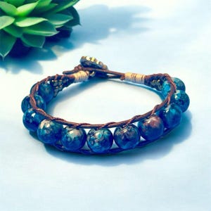May include: A blue beaded bracelet with a brown leather cord. The bracelet features multiple round, blue beads with a marbled appearance. The cord is woven and secured with a gold-colored clasp. The bracelet is a piece of jewelry.