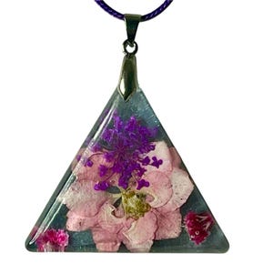 Könnte beinhalten: Eine dreieckige Anhängerkette mit gepressten rosa und lila Blumen, die in klarem Harz eingeschlossen sind. Der Anhänger hängt an einem lila Band und hat eine silberfarbene Metallöse.