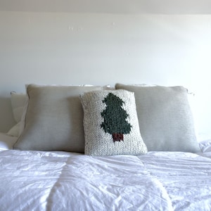 Op de afbeelding: Een wit bed met een wit dekbed en twee lichtgrijze kussens. Een wit kussen met een groen gebreid boomontwerp bevindt zich in het midden van het bed.