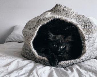 May include: A black cat with green eyes peeks out of a gray and white crocheted cat bed. The bed is on a white bedspread.