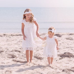 May include: Two girls in white lace dresses walk on a sandy beach. The girl in the front holds a sign that says "Mrs. Daze".