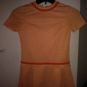 Könnte beinhalten: Orange-weiß kariertes Kleid mit kurzen Ärmeln und einem plissierten Rock. Das Kleid hat einen runden Ausschnitt und einen kontrastierenden orangefarbenen Besatz an Hals und Taille. Es hängt an einem schwarzen Kleiderbügel.